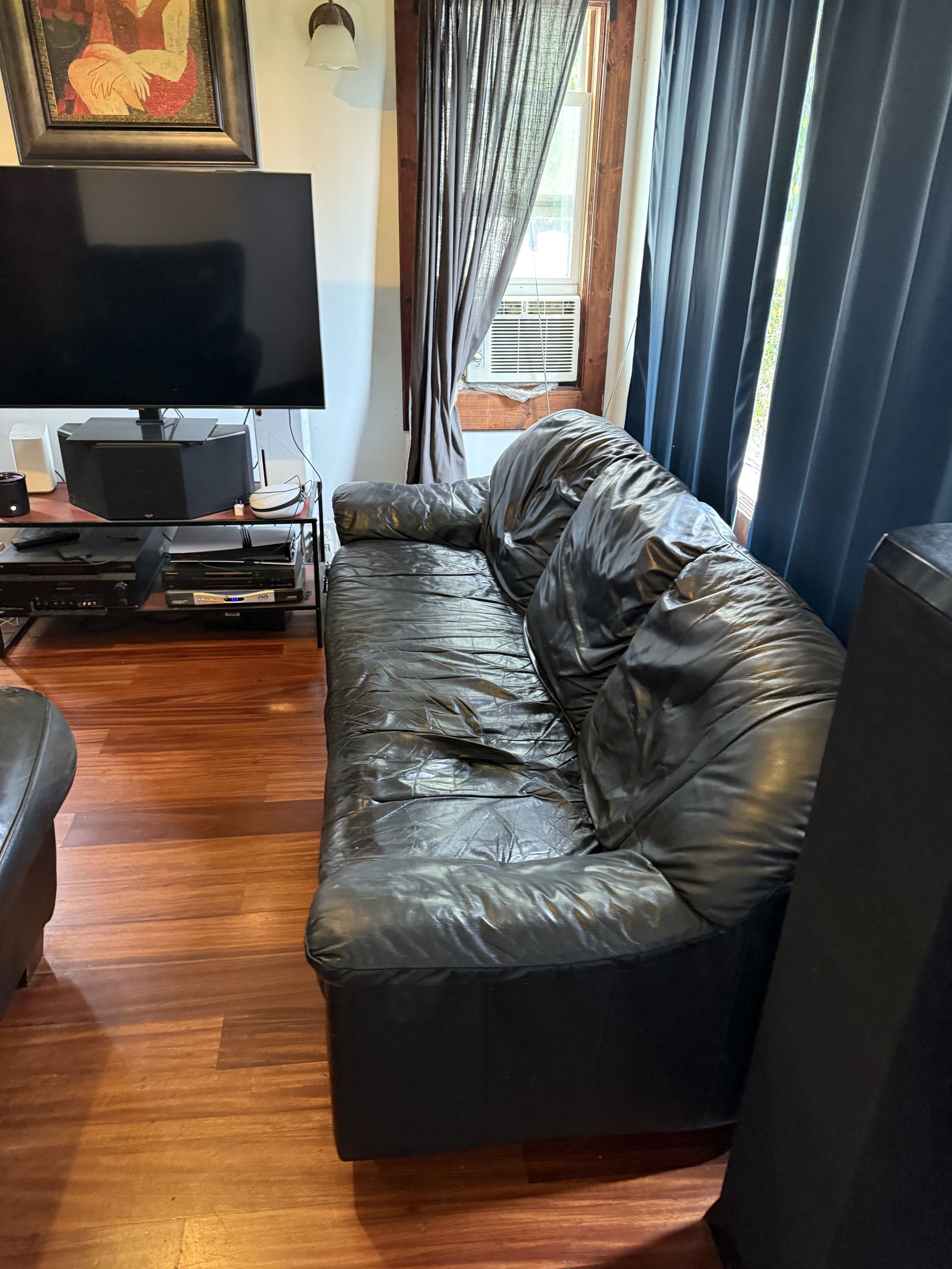 Living room with a black leather couch, TV stand, and dark curtains on hardwood floors