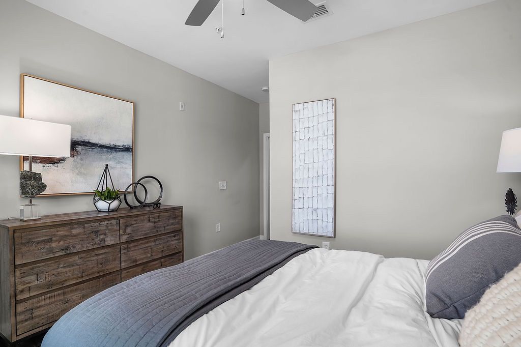Bedroom with a bed, wooden dresser, lamps, and abstract art on neutral walls.