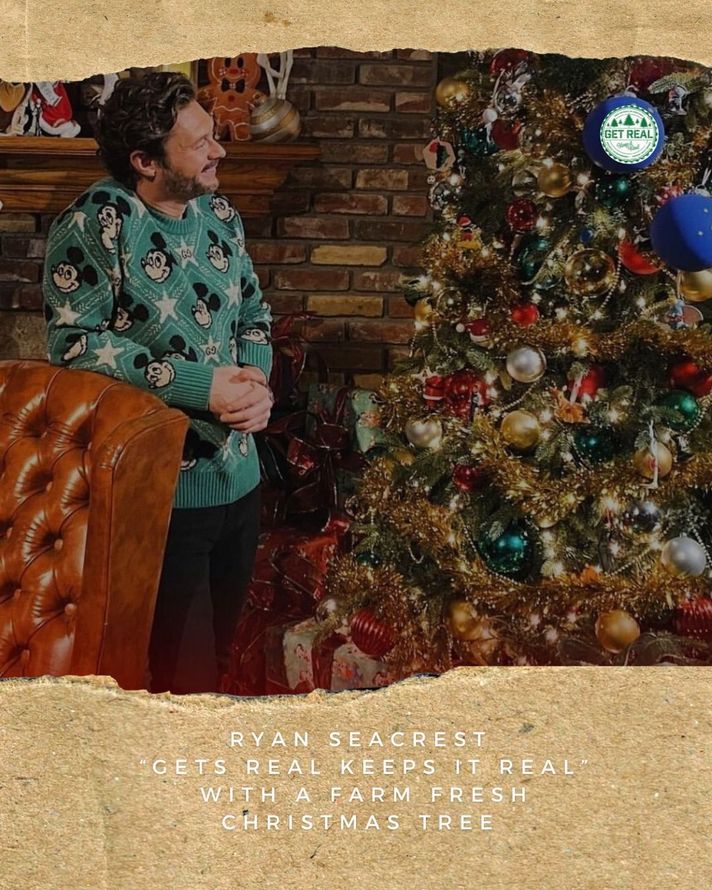 Ryan Seacrest in a sweater is standing in front of a Christmas tree