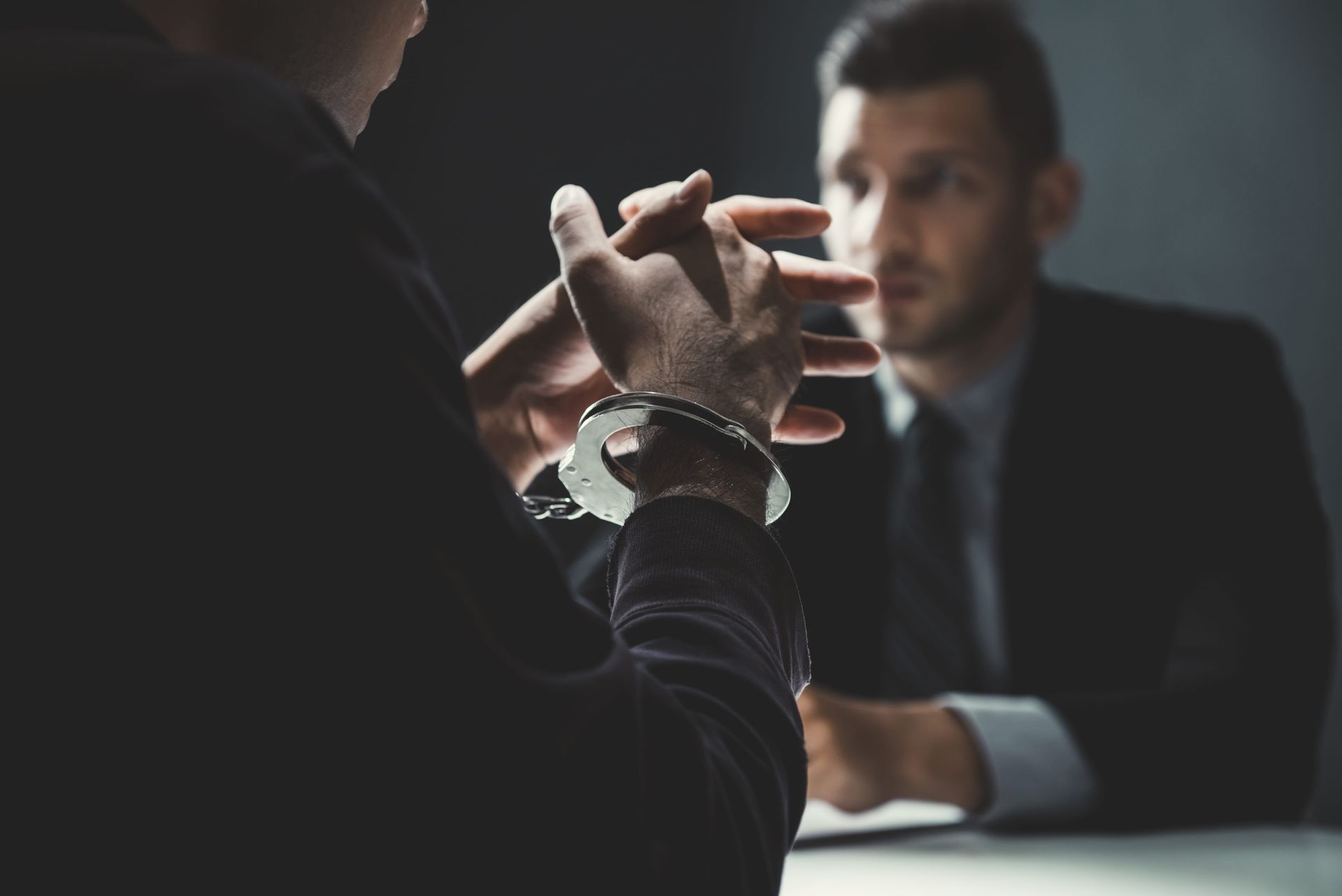 A person wearing handcuffs sits with clasped hands across a table from another person in a dark, professional setting.