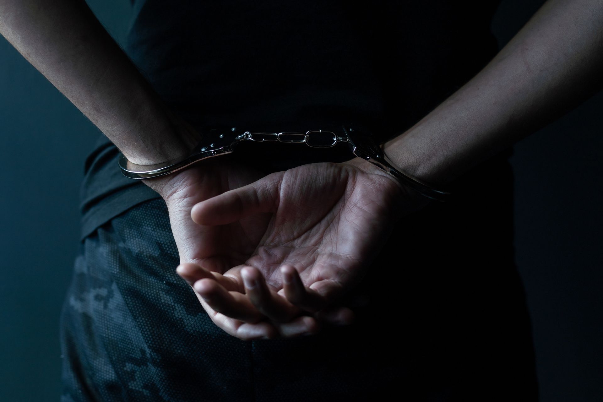 Person in a dark shirt with their hands behind their back in handcuffs against a dark background.
