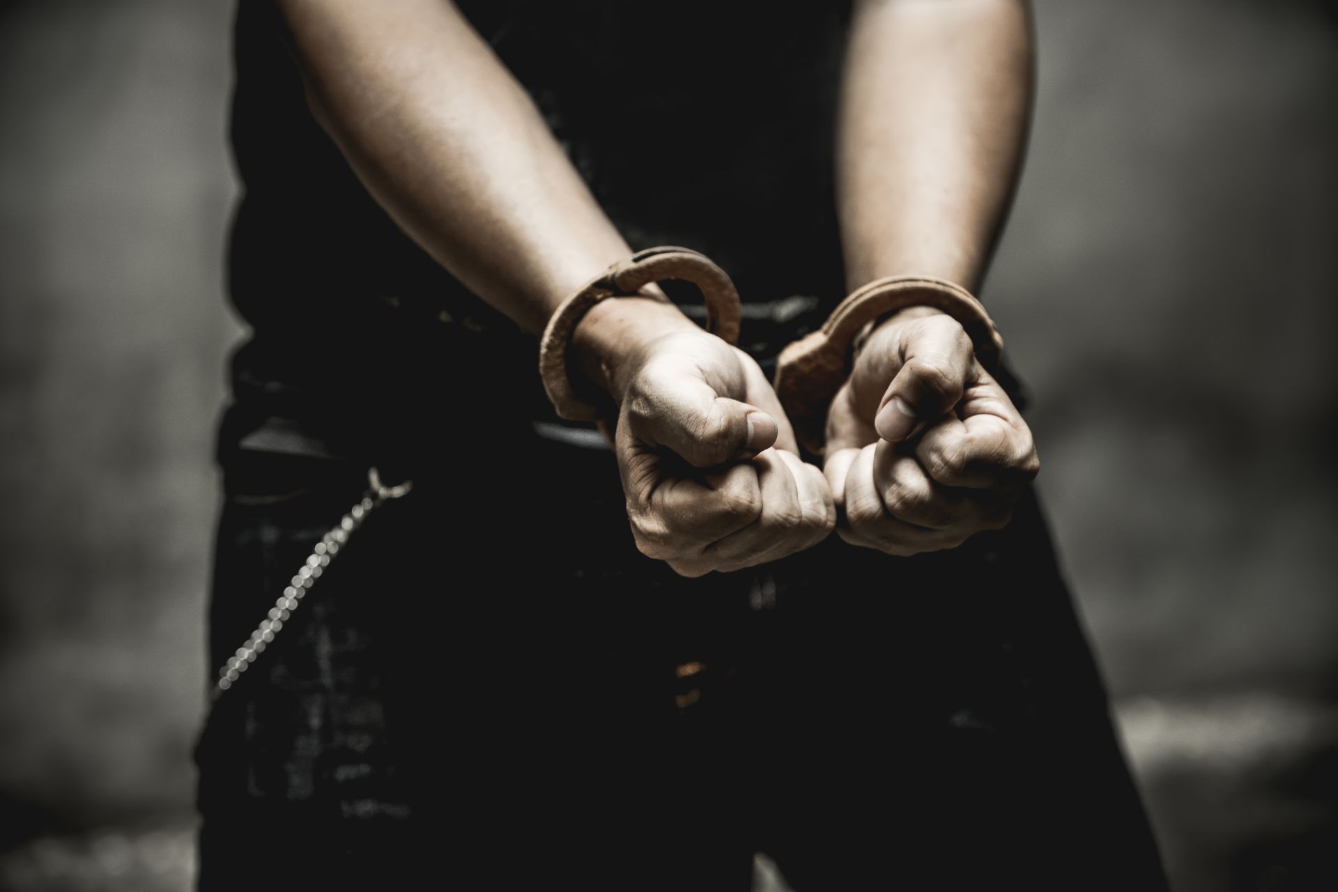 A person in handcuffs; focus on hands. Dark clothing, against a blurred background.