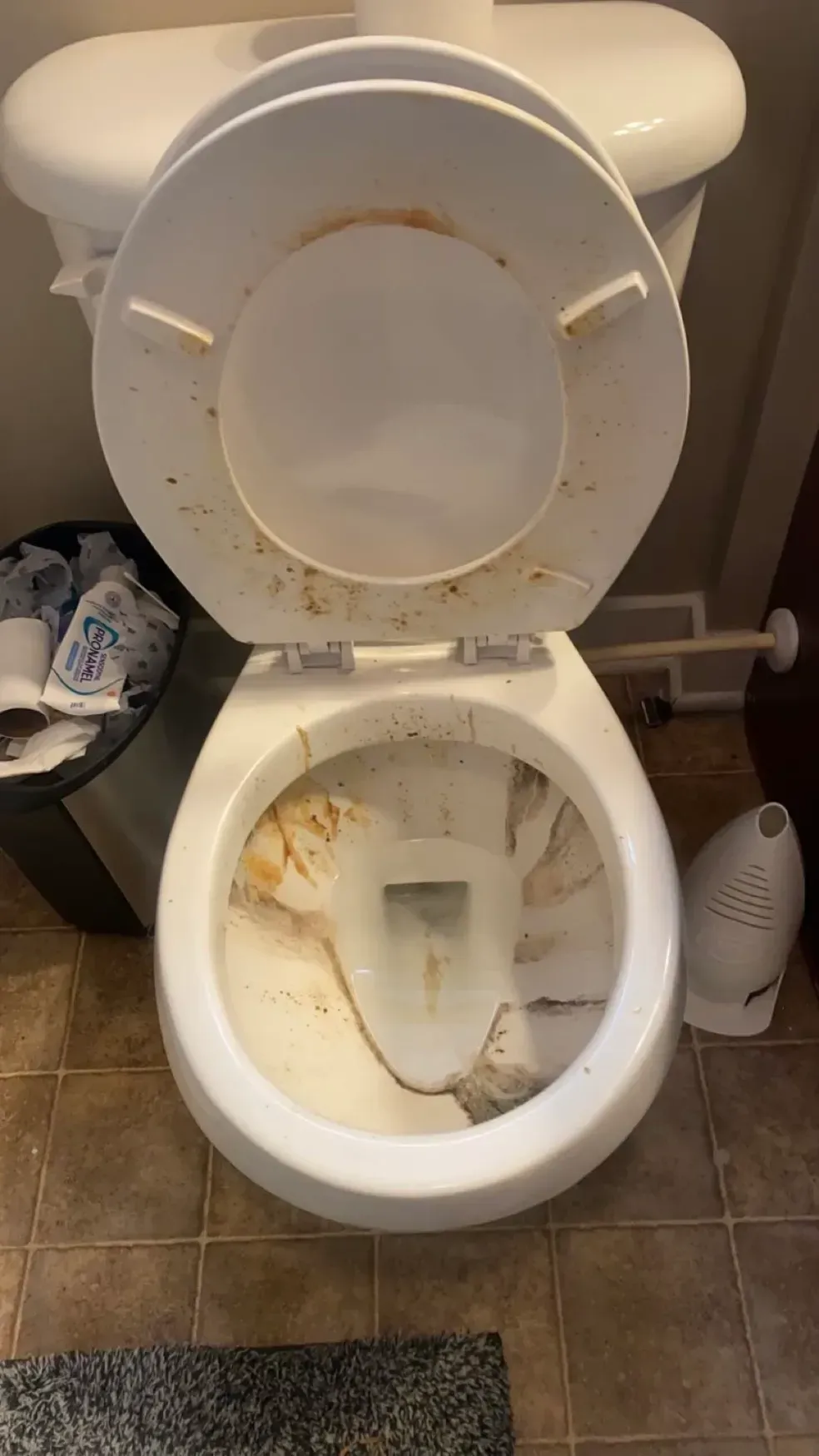 A dirty toilet with the lid up in a bathroom next to a trash can.