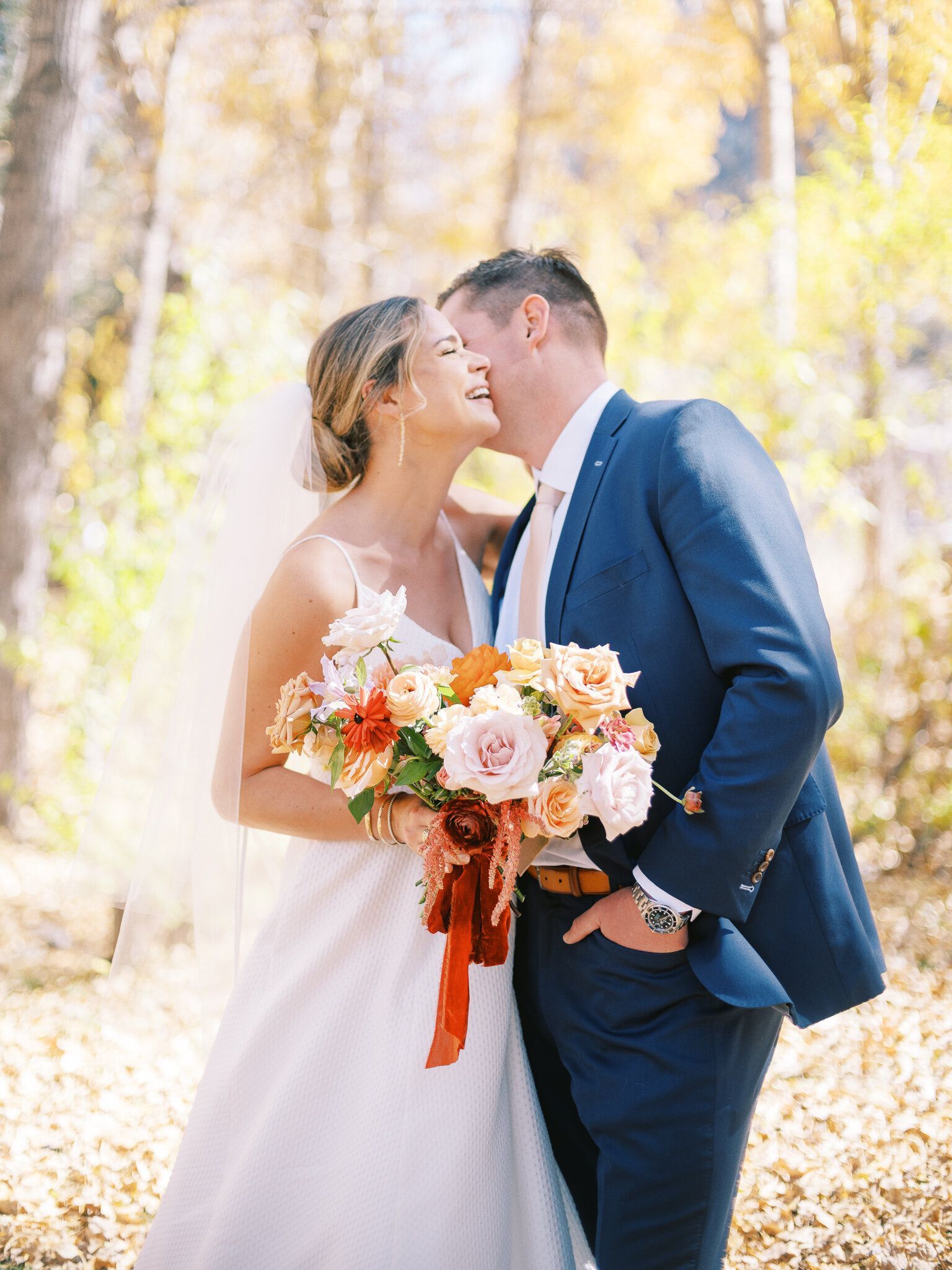 A bride and groom are kissing in the woods while holding a bouquet of flowers.