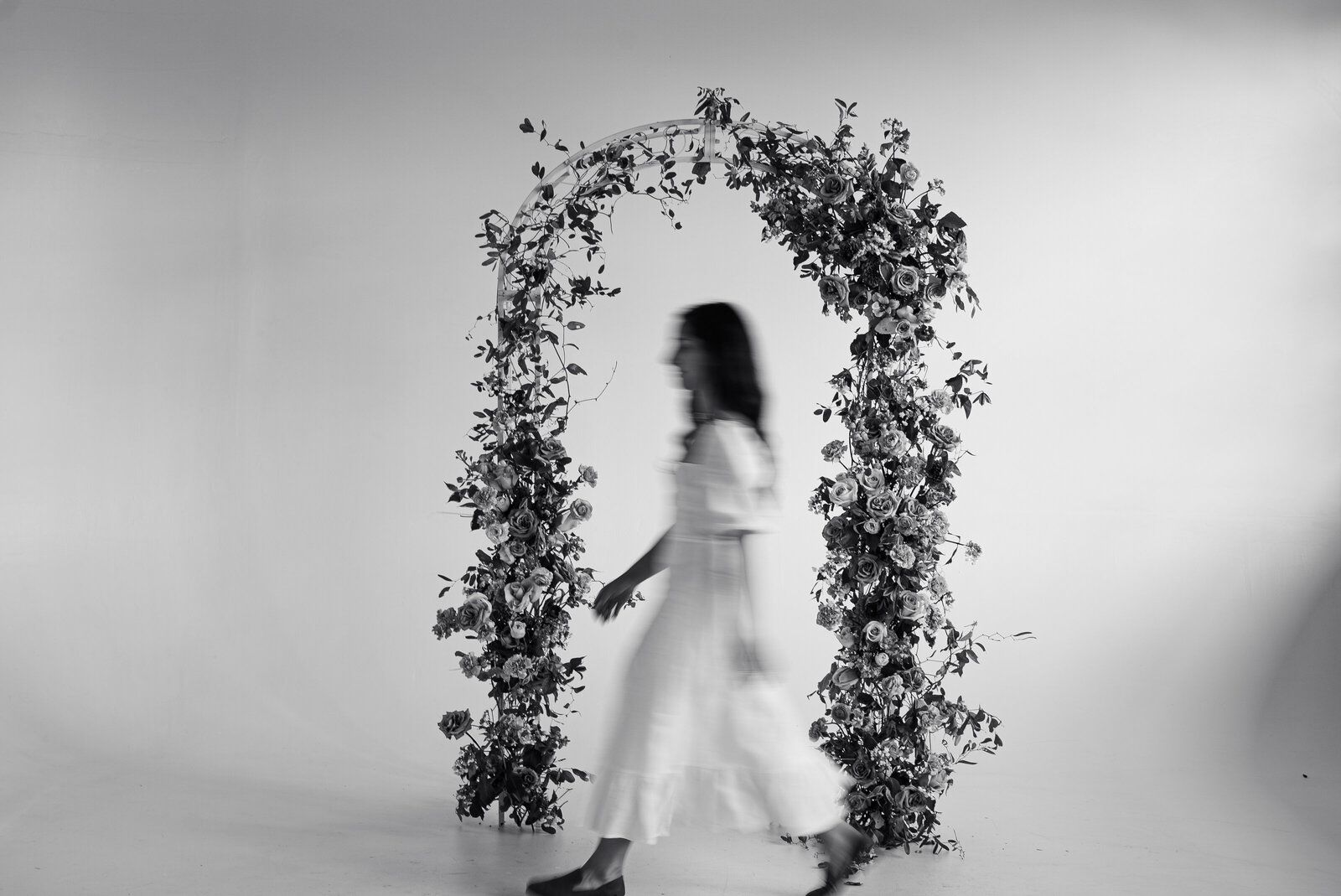 Woman in white dress walking through floral arch. Black and white photo.