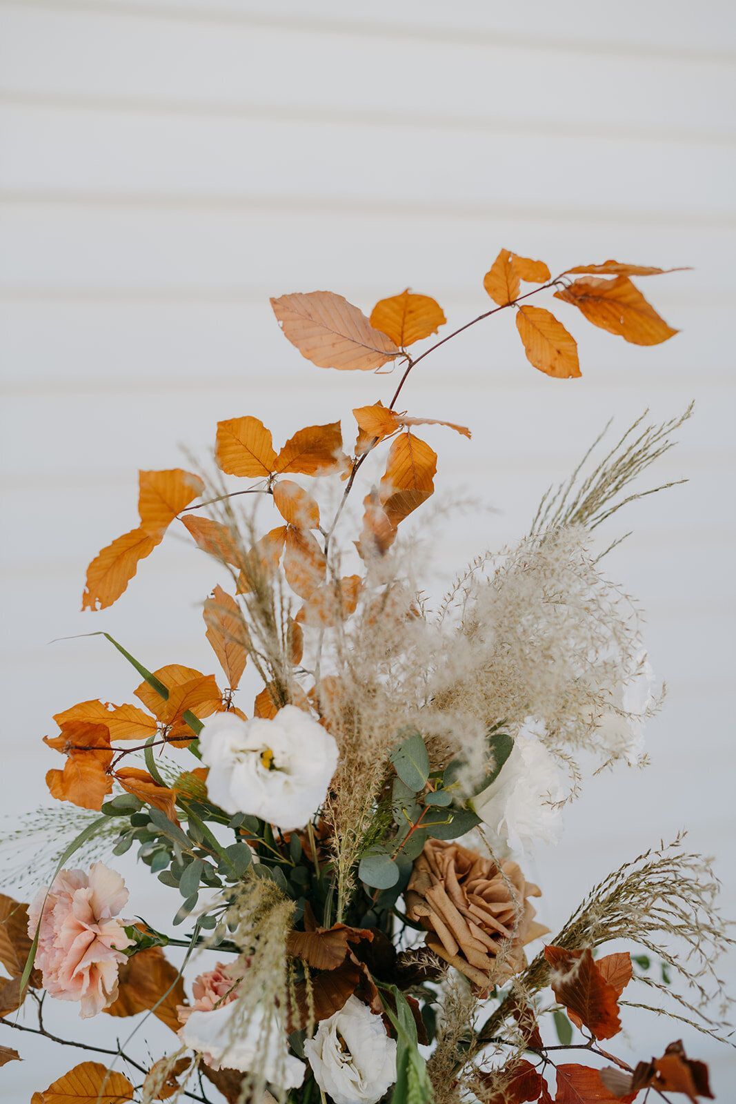 A close up of a vase filled with flowers and leaves against a white wall.