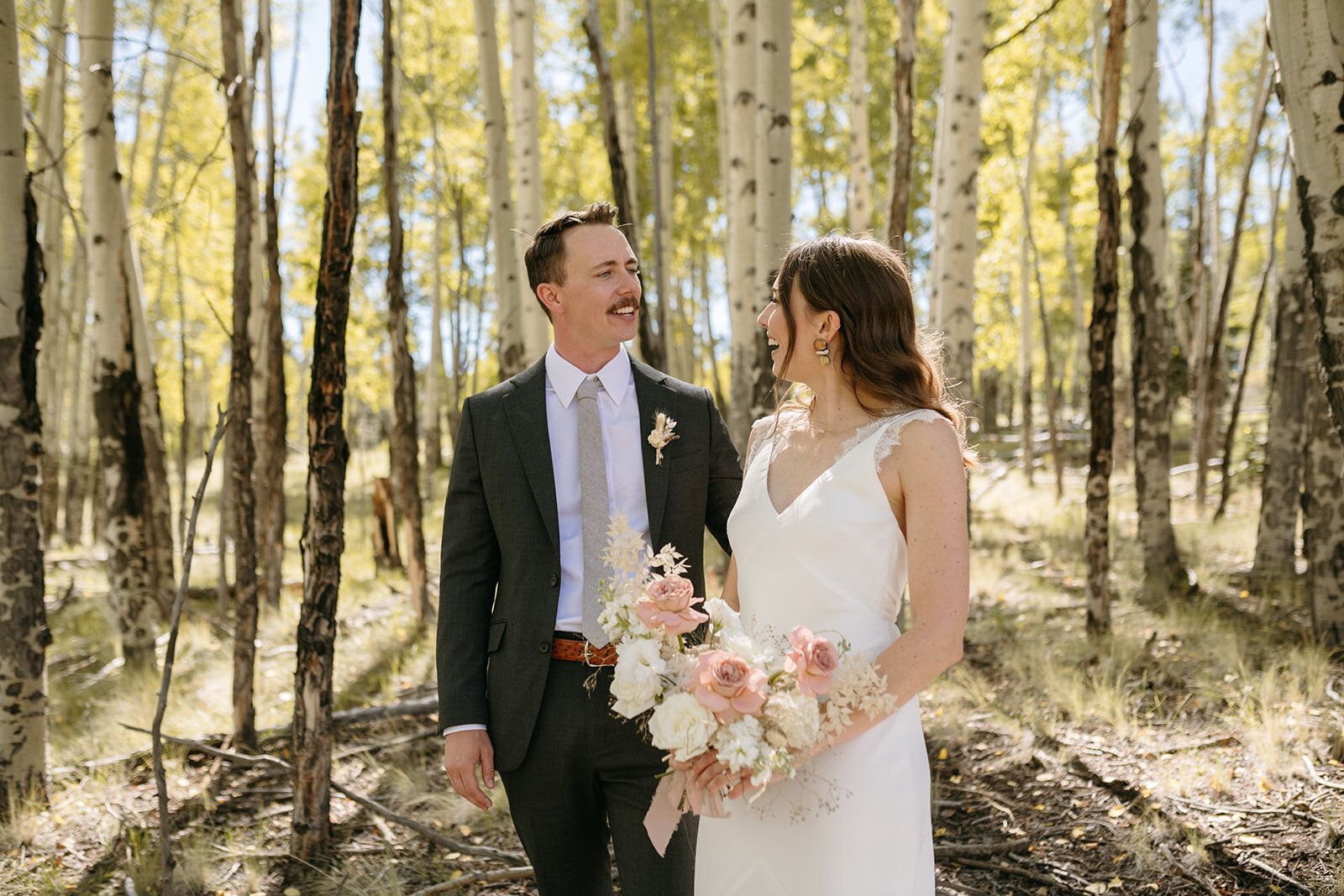 A bride and groom are standing in the woods looking at each other.