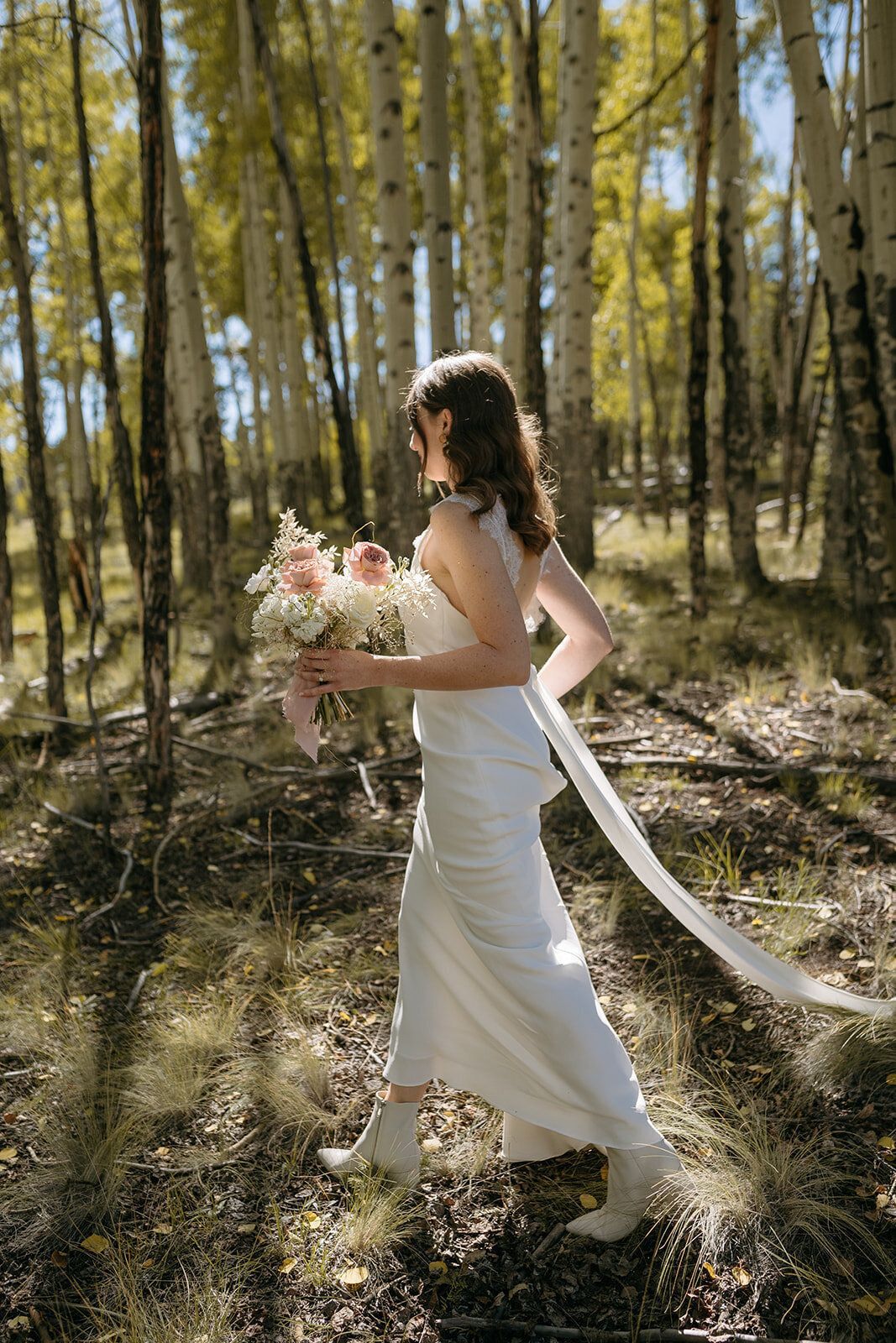 A bride in a white dress is walking through a forest holding a bouquet of flowers.