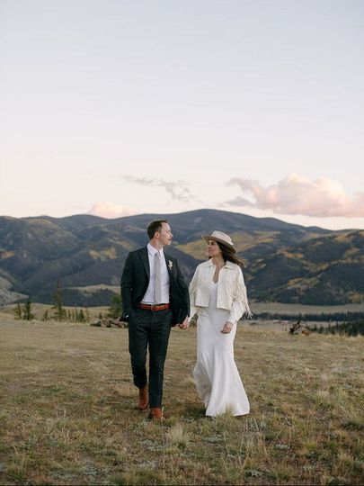 A bride and groom are walking in a field with mountains in the background.