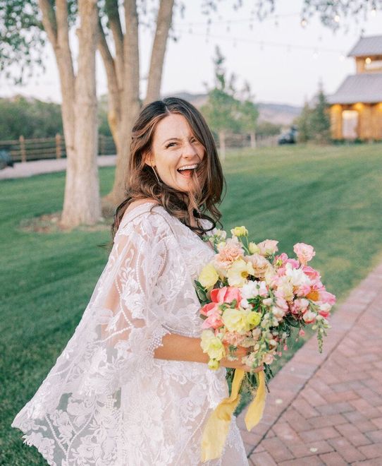 A woman in a white lace dress is holding a bouquet of flowers and smiling.