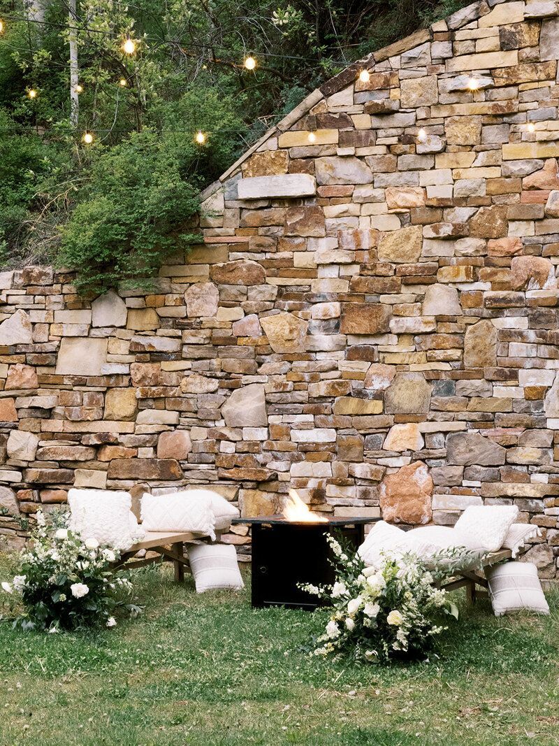 A stone wall with a fire pit and chairs in front of it.