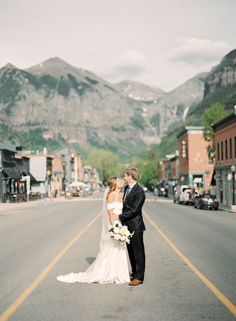 A bride and groom are kissing in the middle of a street with mountains in the background.