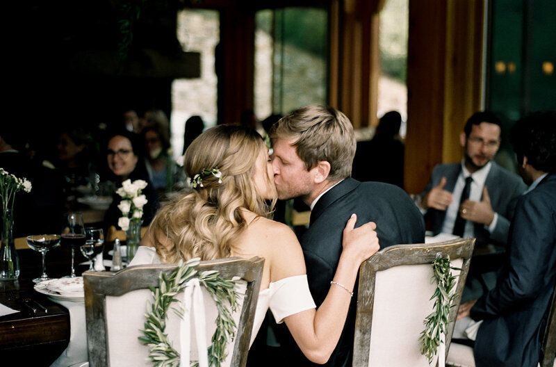 A bride and groom are kissing while sitting in chairs at a wedding reception.