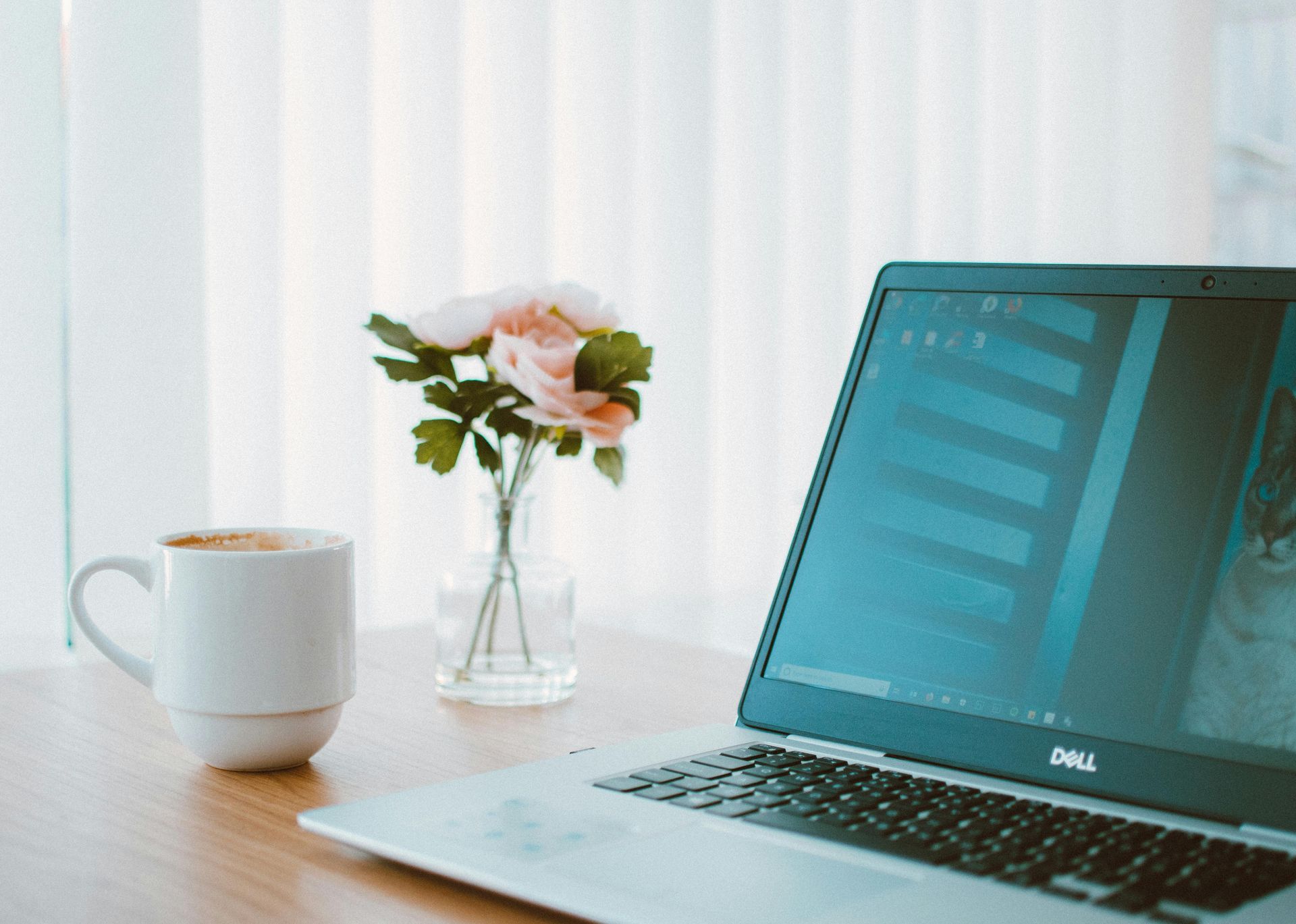 Laptop and coffee cup on a desk by a window with flowers.