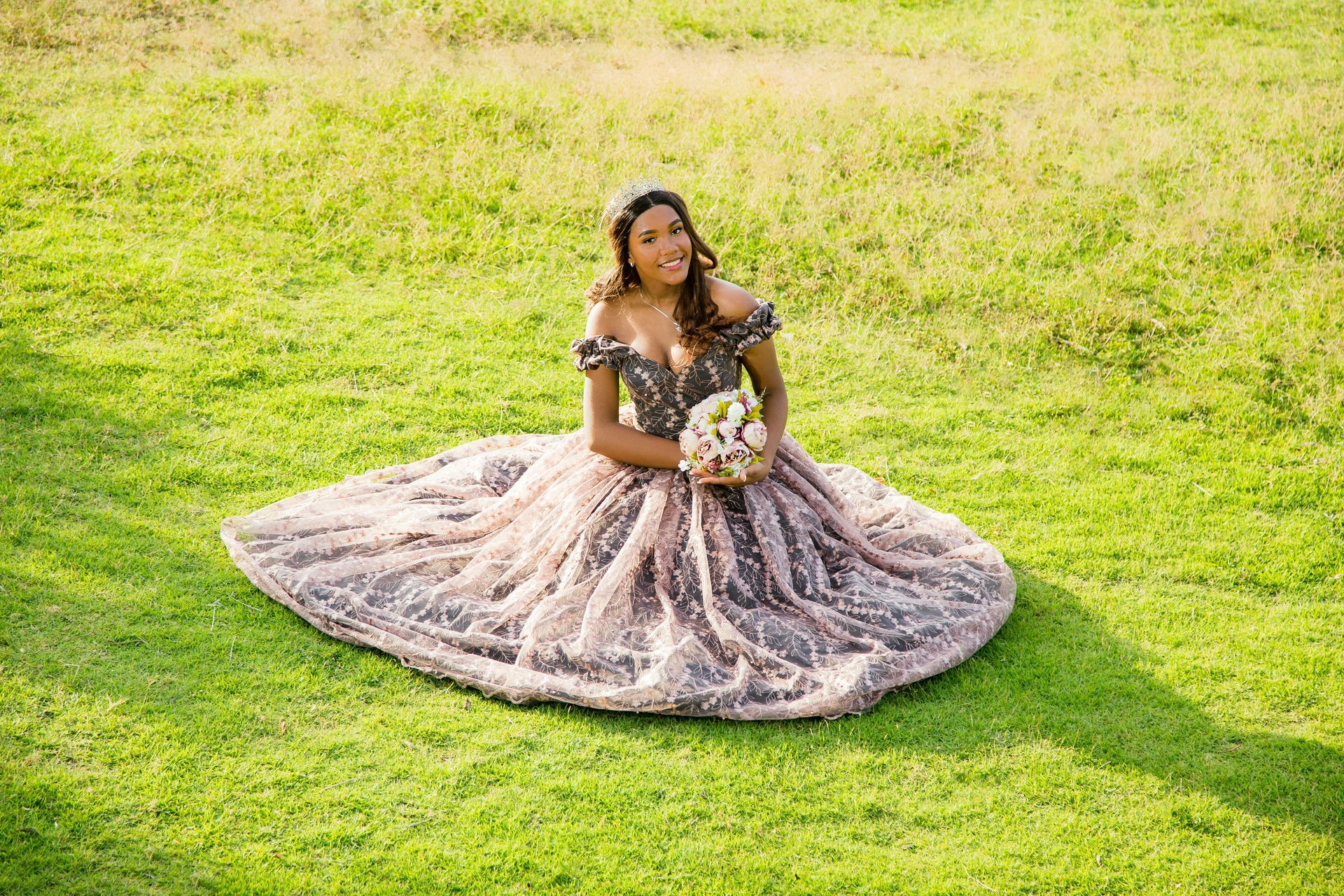 A woman in a long dress is sitting in the grass holding a bouquet of flowers.