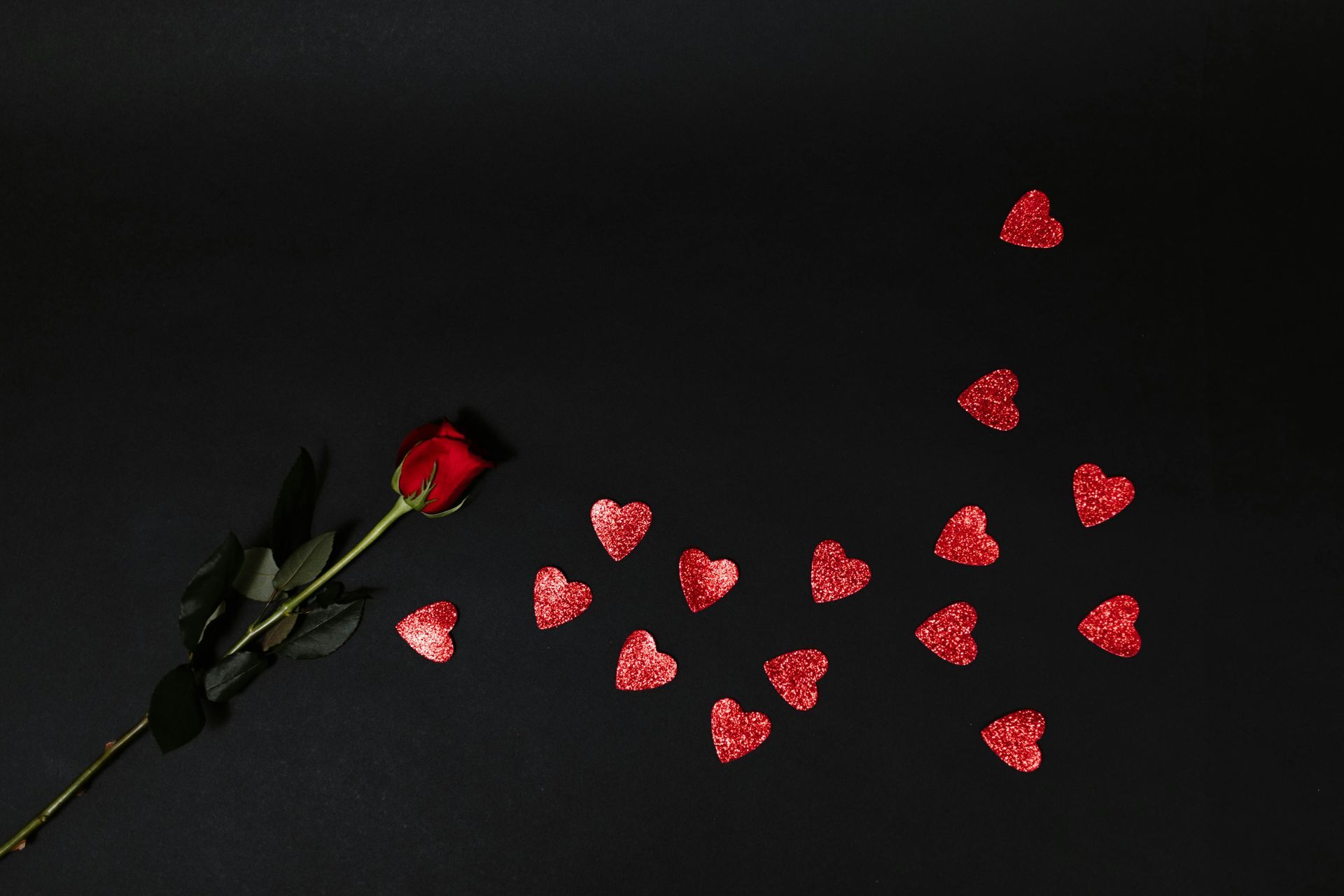 Red rose with a trail of red heart confetti on a black background.