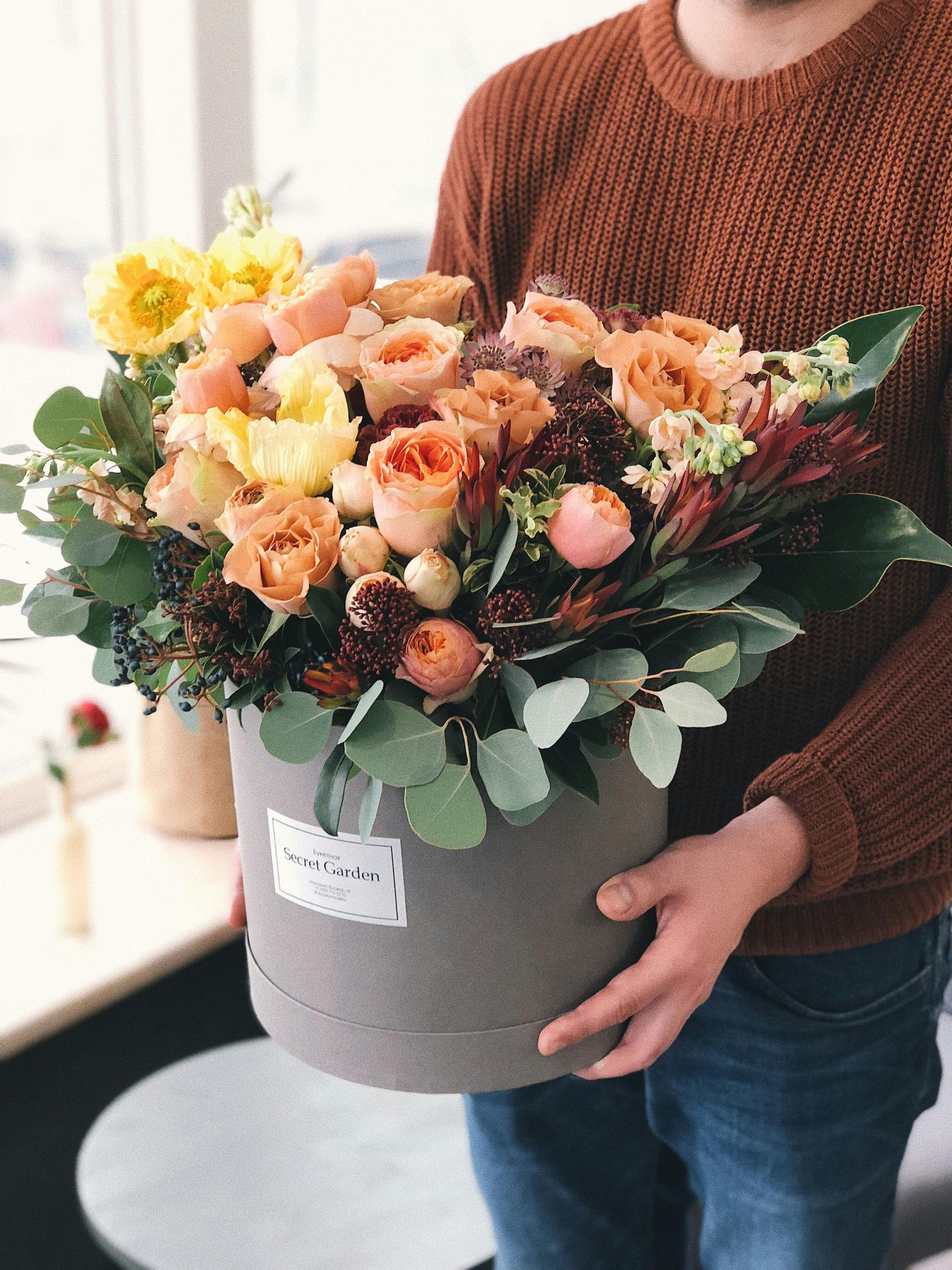 Person holding a large round gray hatbox overflowing with colorful flowers, including orange, yellow, and peach roses, greenery, and berries.