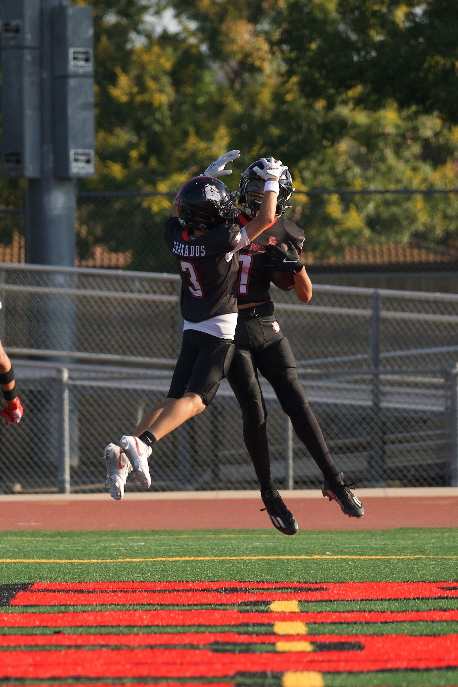 Football players in action on a grassy field; one runs with the ball, pursued by two opponents.