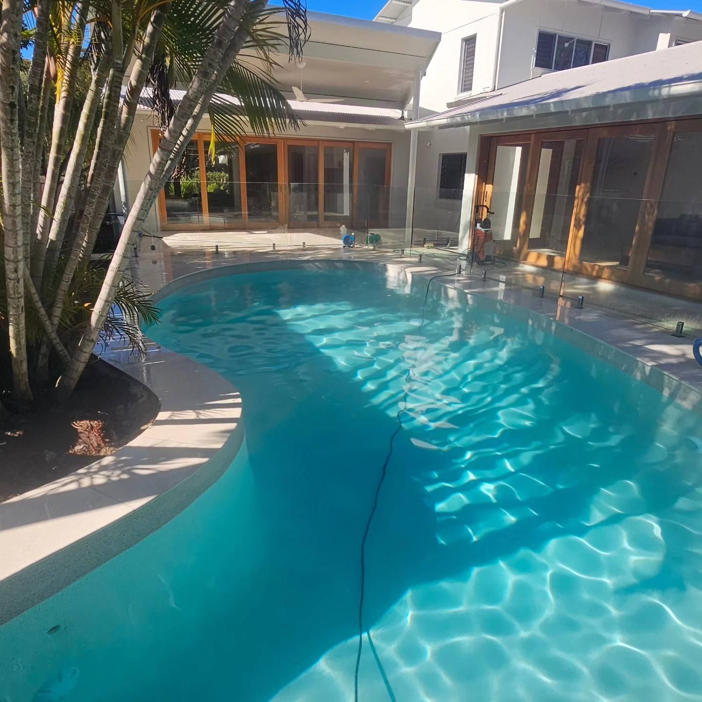 Swimming pool with clear blue water in front of a white house with wooden doors and a glass fence — Sunny Tiling Co in Mount Coolum, QLD