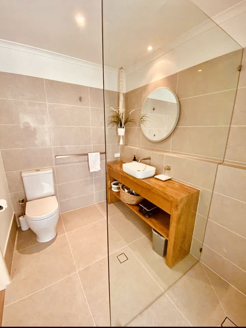 Bathroom with a toilet, wooden vanity, and glass shower. Beige tile, a round mirror, and hanging plant.