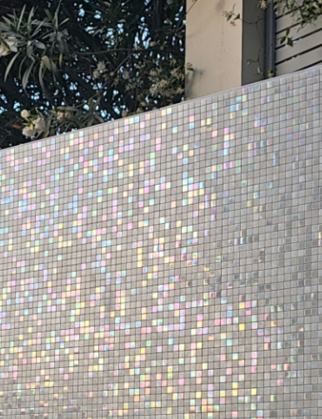 Iridescent mosaic tile wall with dappled sunlight, trees and a building exterior.