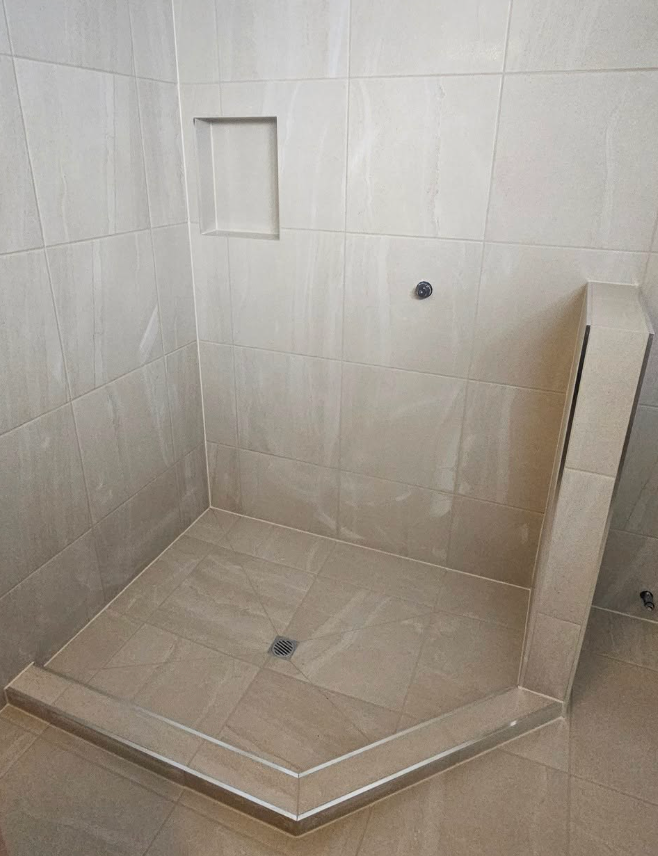 A tiled, beige corner shower stall with a built-in shelf and drainage hole.