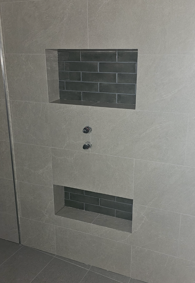Gray tiled shower with two recessed shelves tiled with dark gray brick, and chrome shower controls.