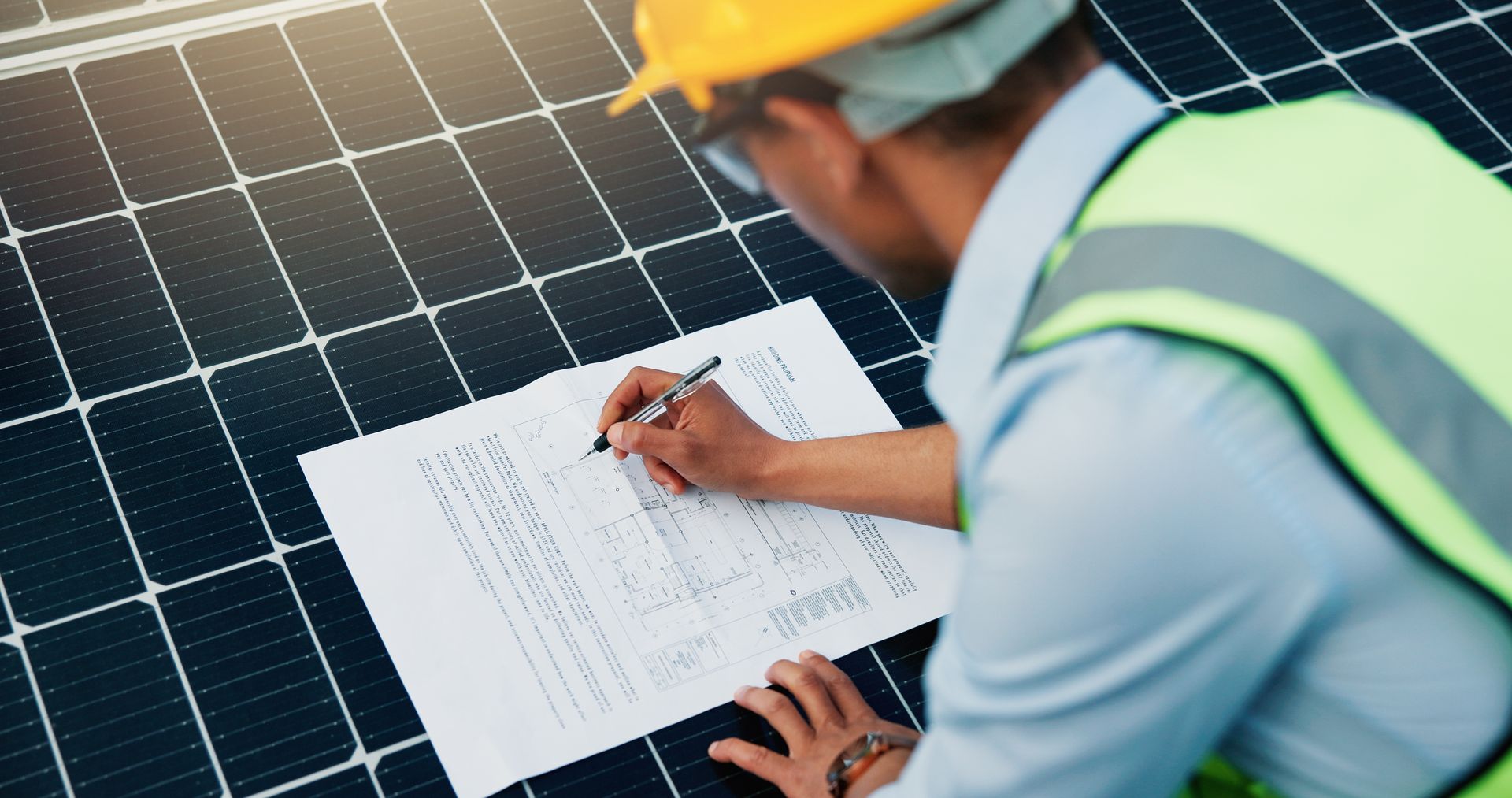 Person in hard hat and safety vest, writing on a document placed on solar panels.