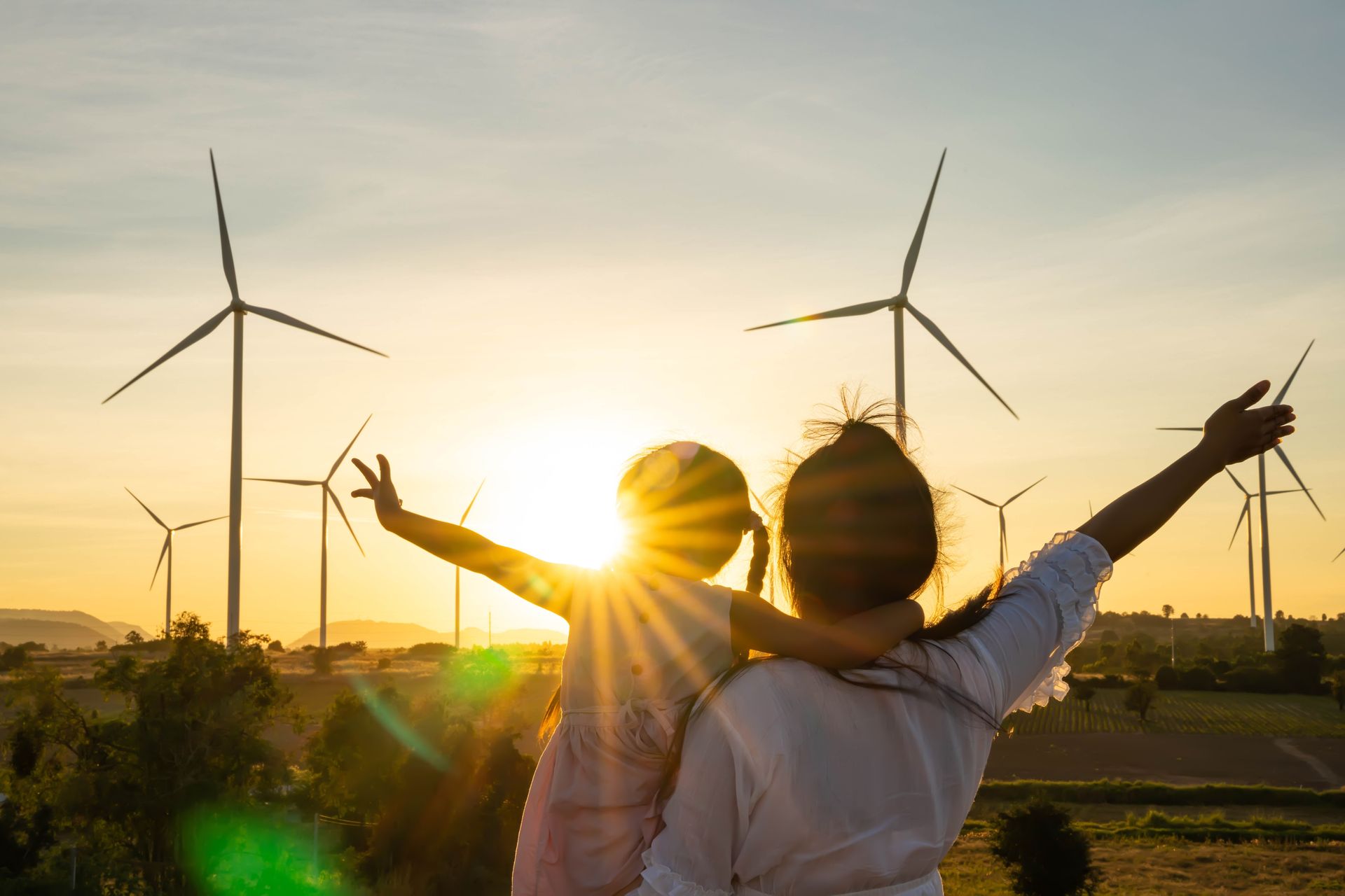 Woman and child with arms raised, looking at a sunset with wind turbines in the background.