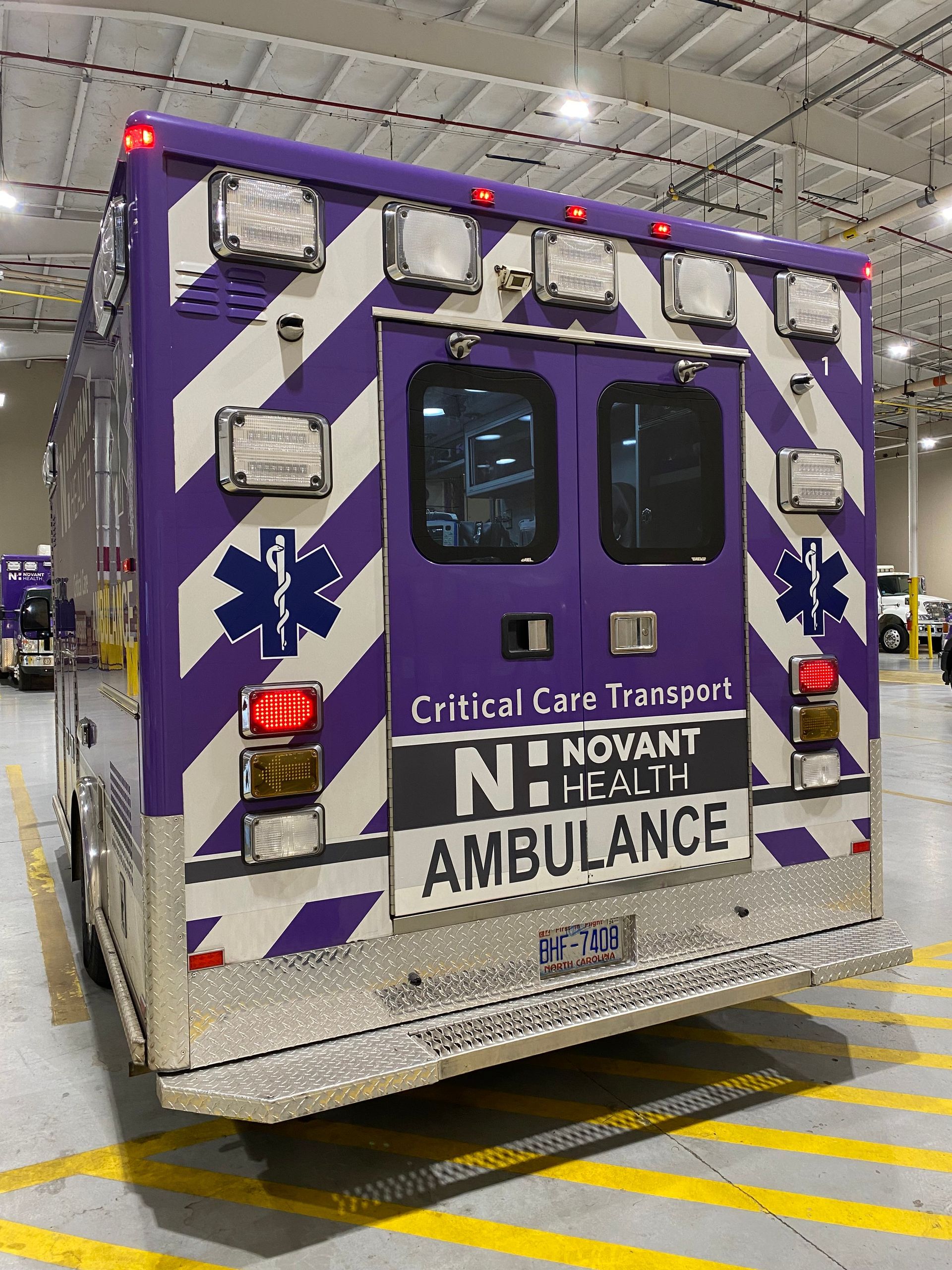 Rear view of a purple-and-white ambulance with flashing lights in a garage, labeled “Critical Care Transport”