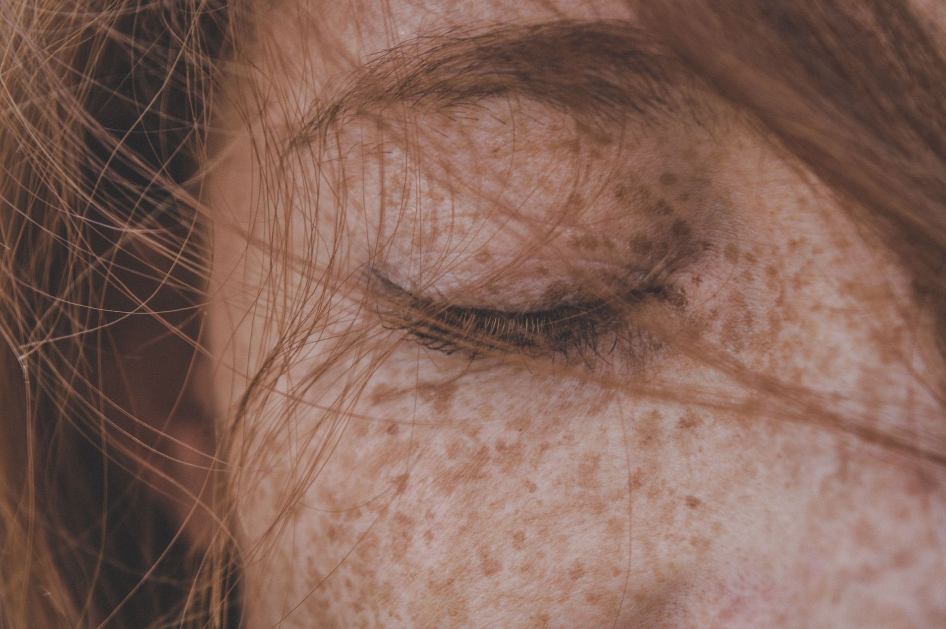 Close-up of a freckled face with a closed eye and strands of hair across it.
