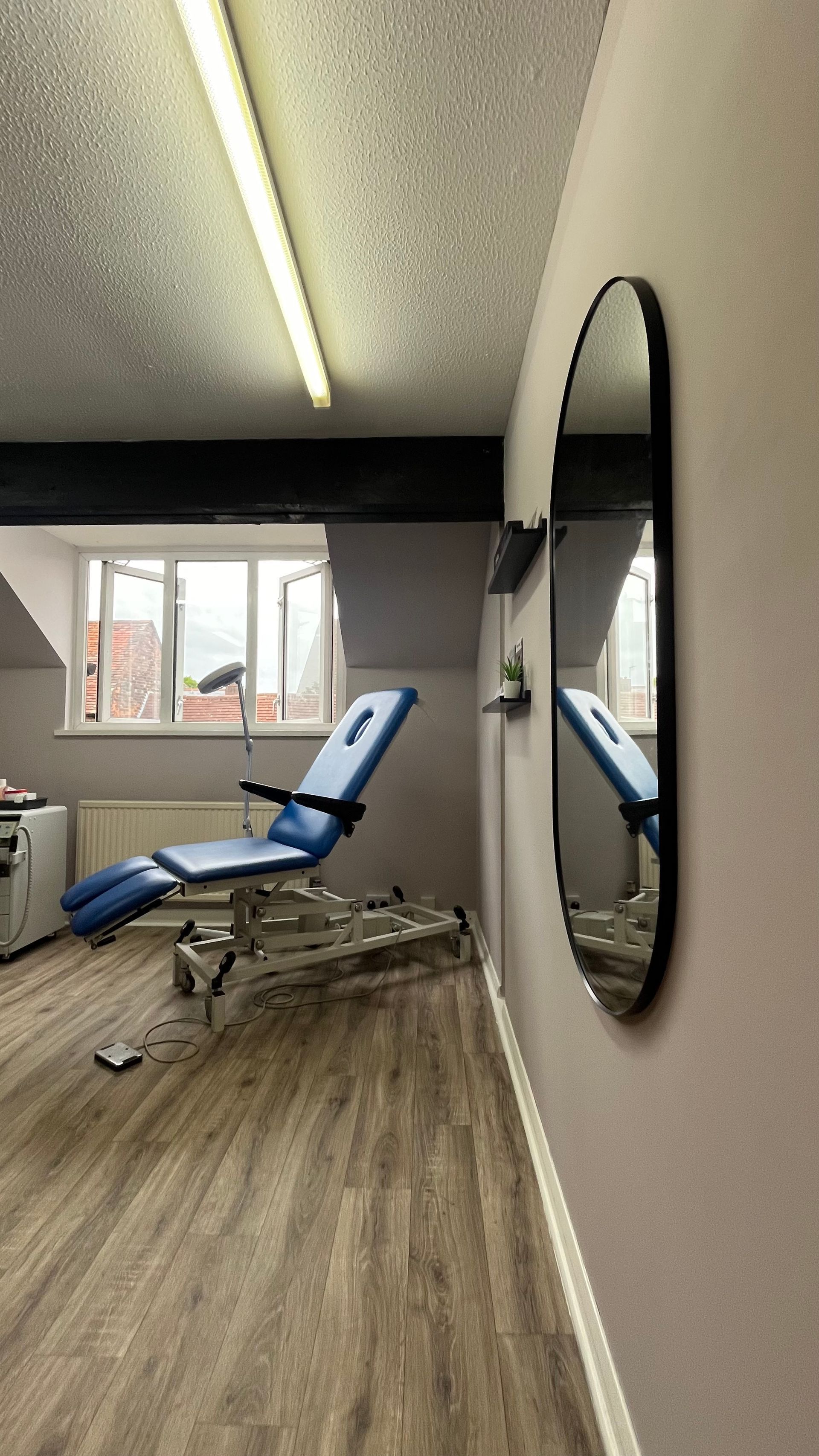Treatment room with a blue chair, oval mirror, and wood-look floor.