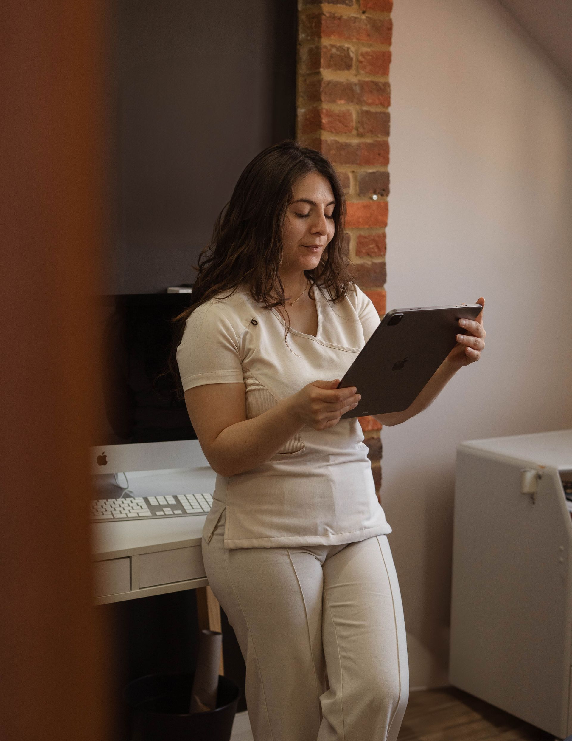 Woman in cream coloured scrubs leaning against brick wall, using a tablet.
