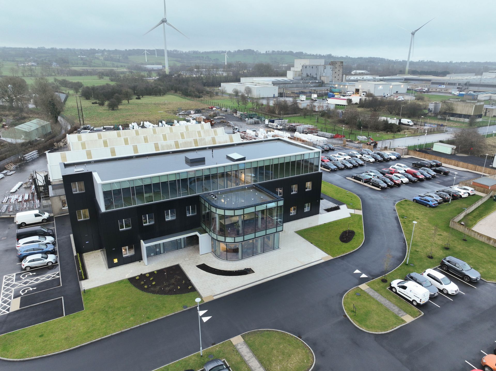 Modern office building with glass facade and cars parked in front; windmills in background.
