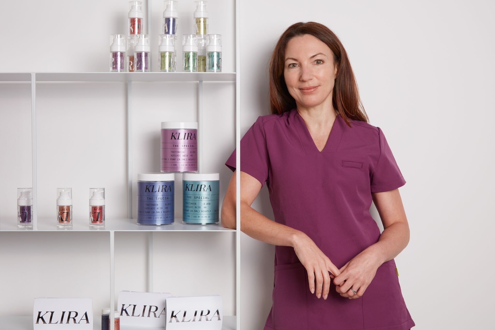 Woman in purple scrubs stands by a shelf of cosmetic products, smiling.
