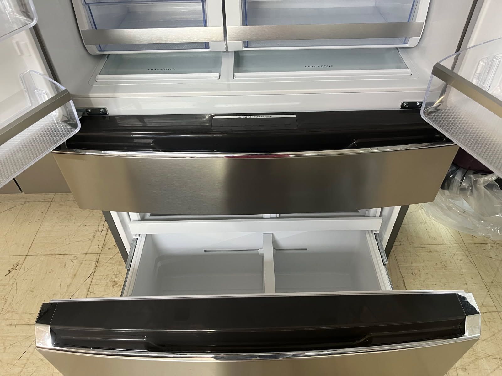 A refrigerator with an open lower freezer drawer and a shelf above, showing a glossy black and stainless steel interior.