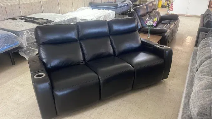 Black leather reclining loveseat in a furniture showroom, with beds and other sofas in the background.