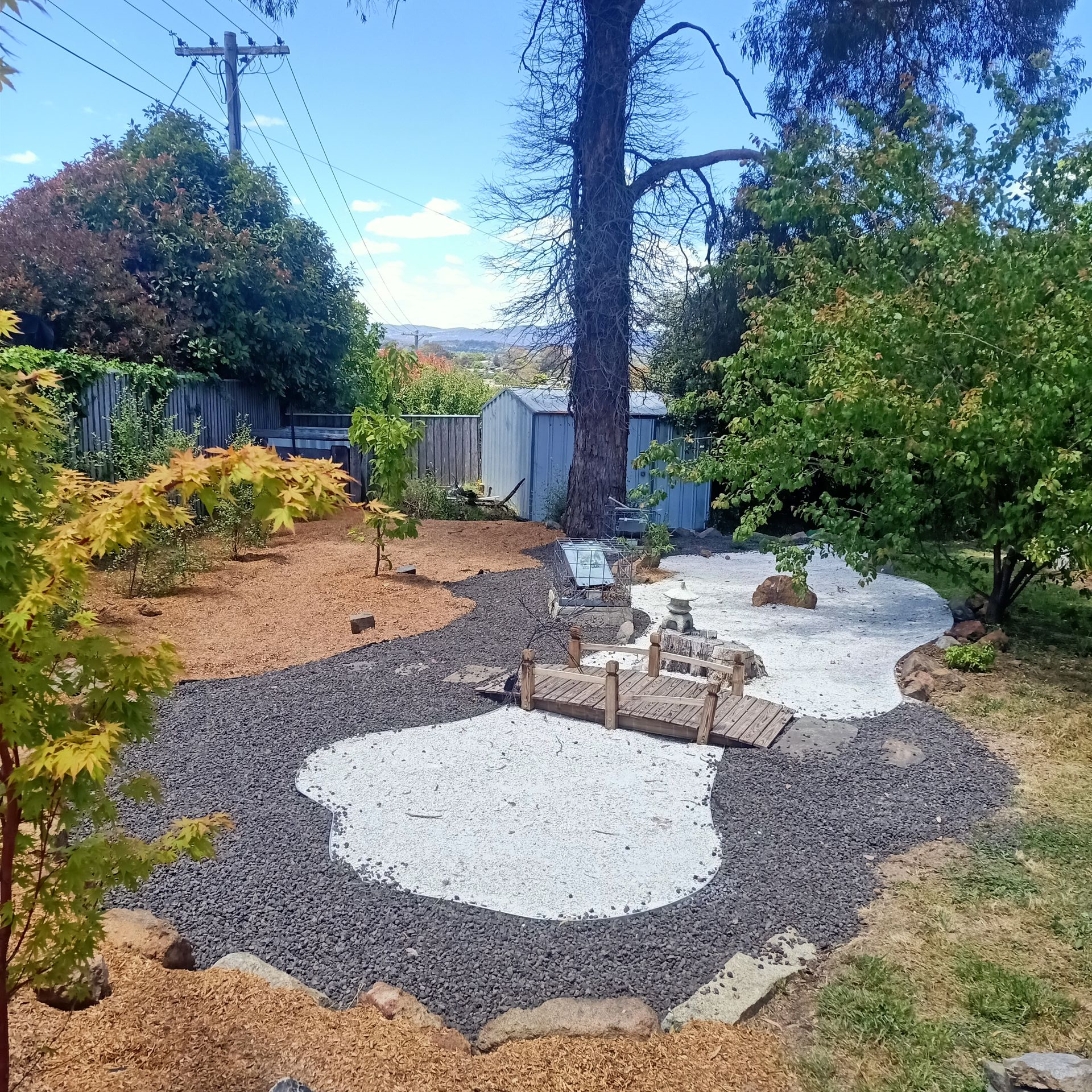 A decorative garden with a gravel path, white stone area, and small wooden bridges, surrounded by trees. — Your Garden in Charnwood, ACT