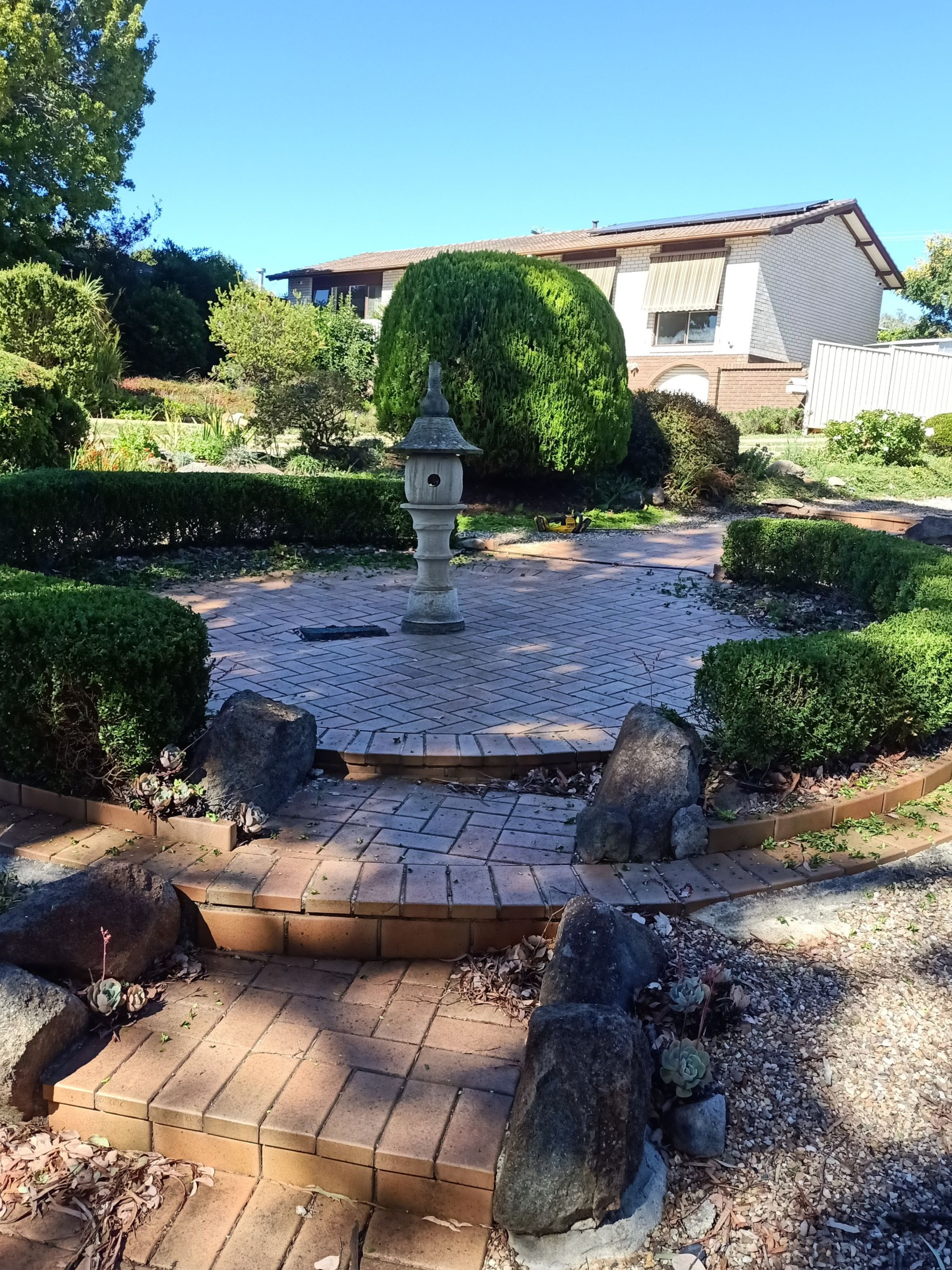 A Japanese garden with stone path, lantern, and trimmed bushes, under a blue sky. — Your Garden in Charnwood, ACT