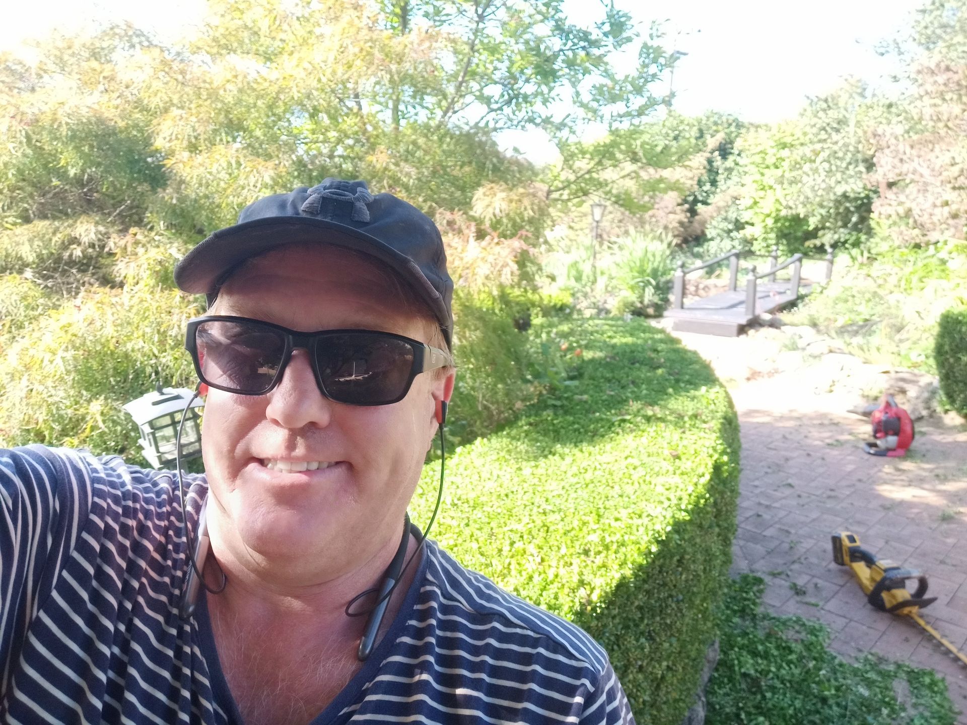 A smiling person wearing a striped shirt, sunglasses, and a cap outdoors, with a garden hedge and lawn tools nearby. — Your Garden in Gungahlin, ACT