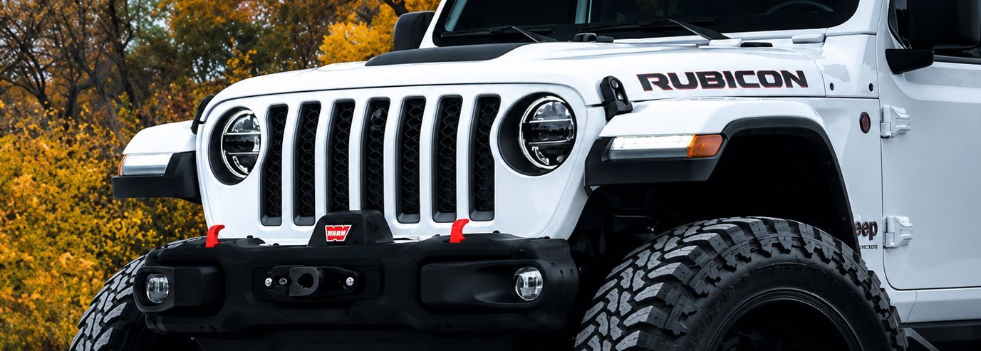 White Jeep Rubicon front view with off-road tires, parked outdoors against autumn trees.