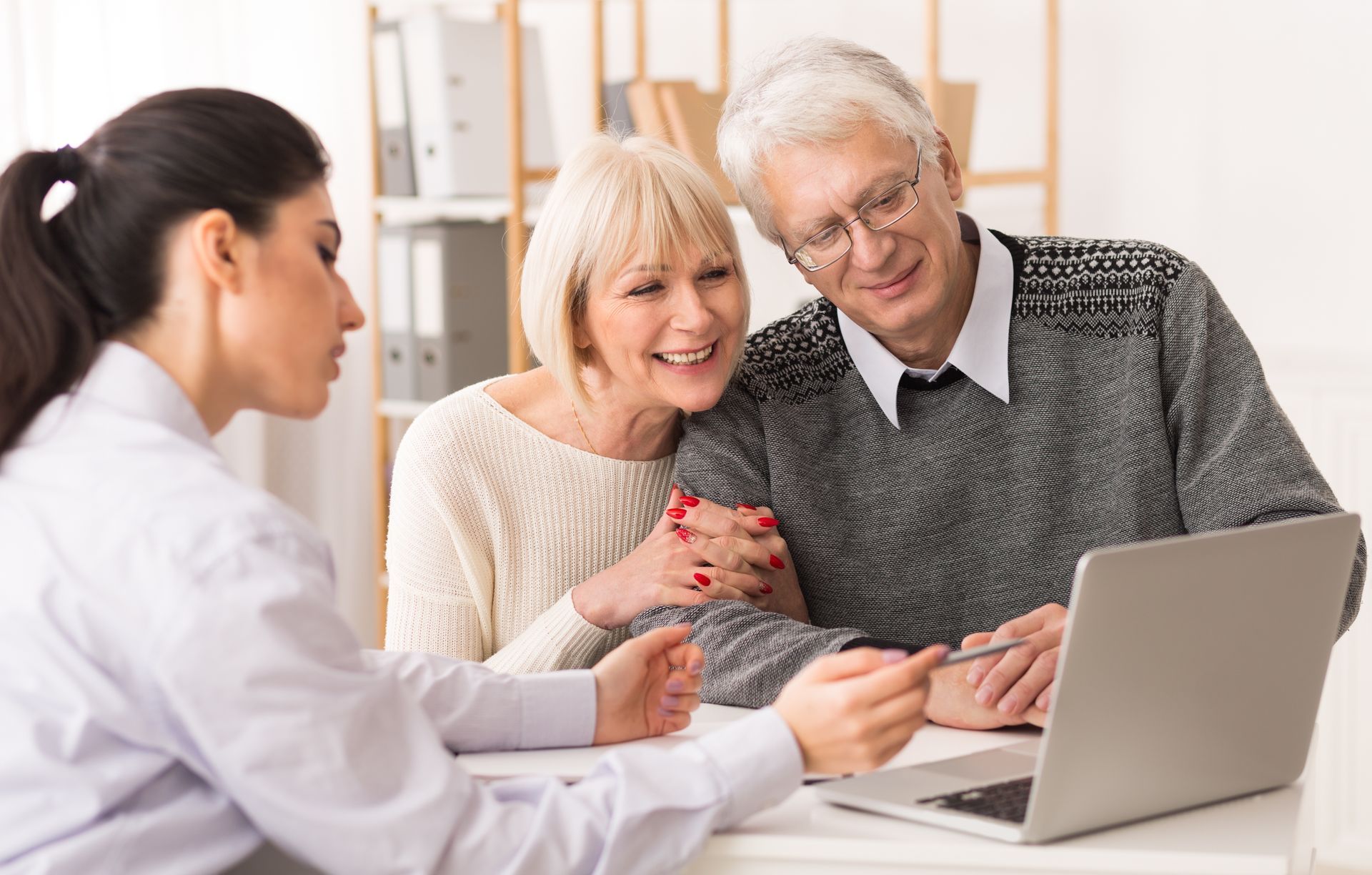 Estate Planning advisor pointing at laptop screen, smiling at elderly couple in office.