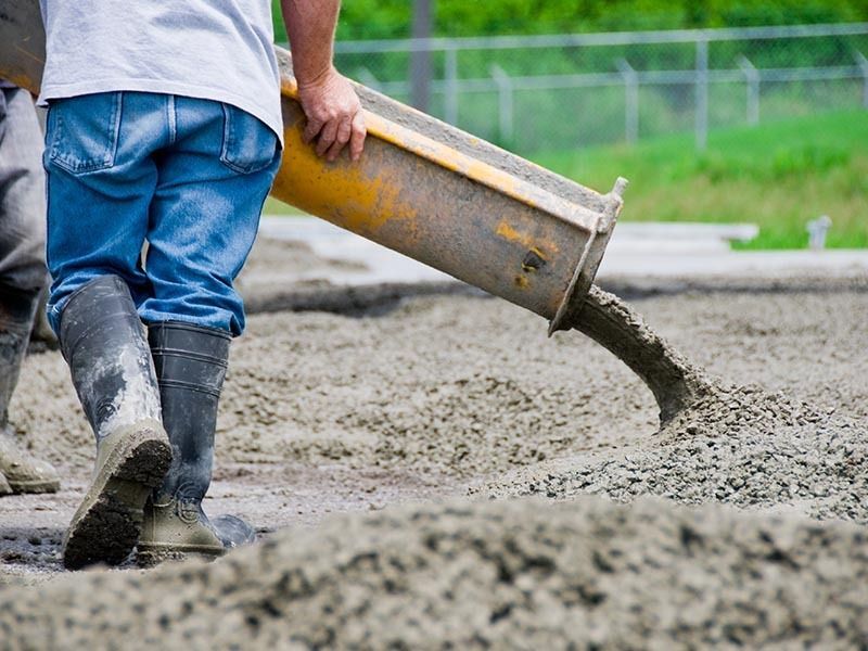 A construction worker in blue jeans and rubber boots directs wet concrete from a yellow chute onto a site.