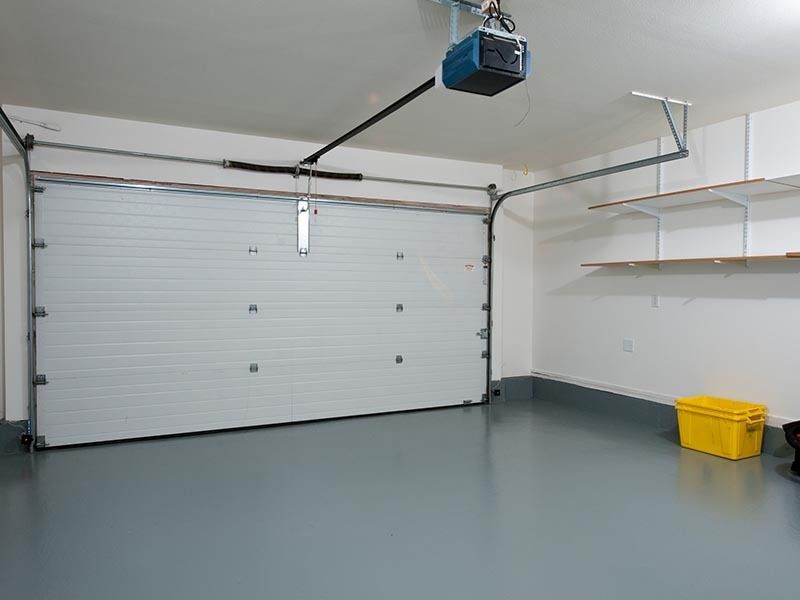 A clean, modern garage featuring a closed white sectional door, gray epoxy flooring, wall shelving, and a yellow bin.