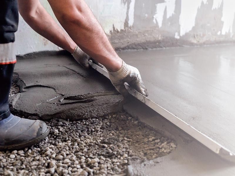 A worker uses a long straightedge tool to level wet concrete over a gravel base on an indoor construction site.