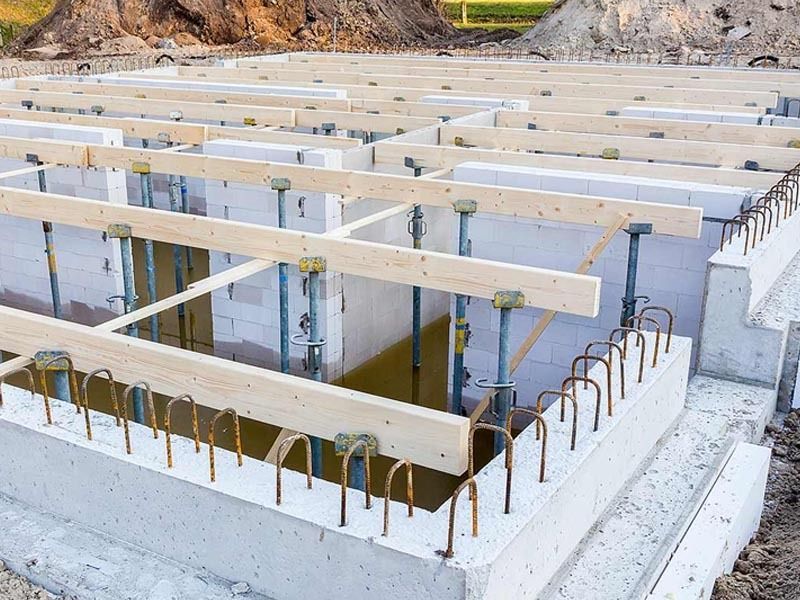 Construction site featuring a concrete foundation with upright steel supports and wooden beams for a flooring system.