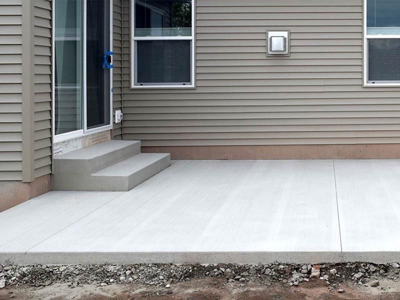 A newly poured concrete patio with two steps leading to a sliding glass door on a house with beige vinyl siding.