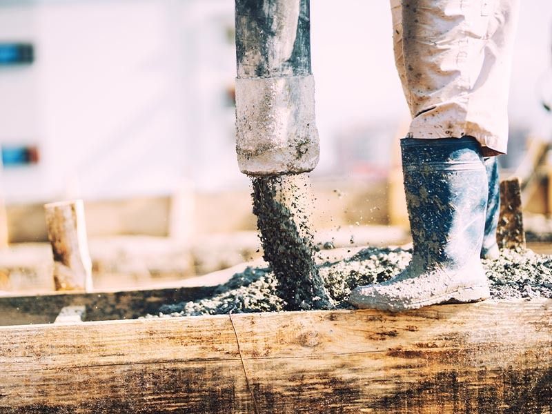 A construction worker in rubber boots pours concrete into a wooden form on a building site.