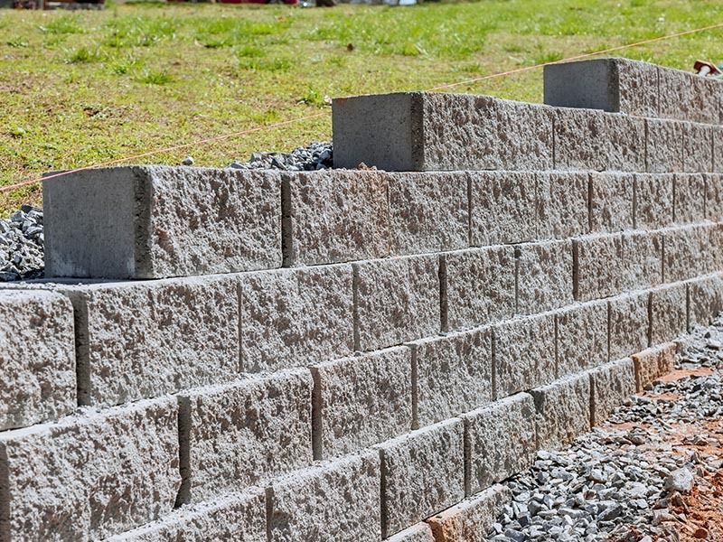 A low retaining wall made of rough, grey stone blocks, partially built into a grassy slope with loose gravel at the base.
