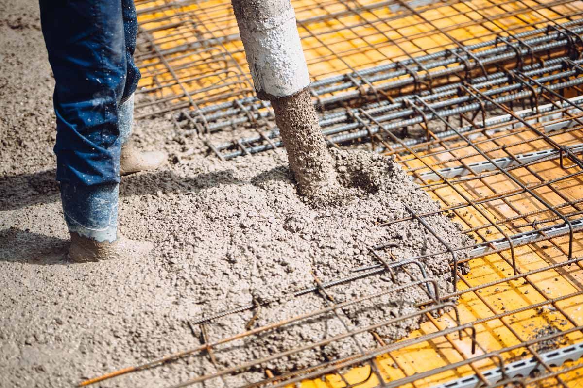A worker in boots pours wet concrete from a hose onto a metal rebar grid atop a yellow construction surface.